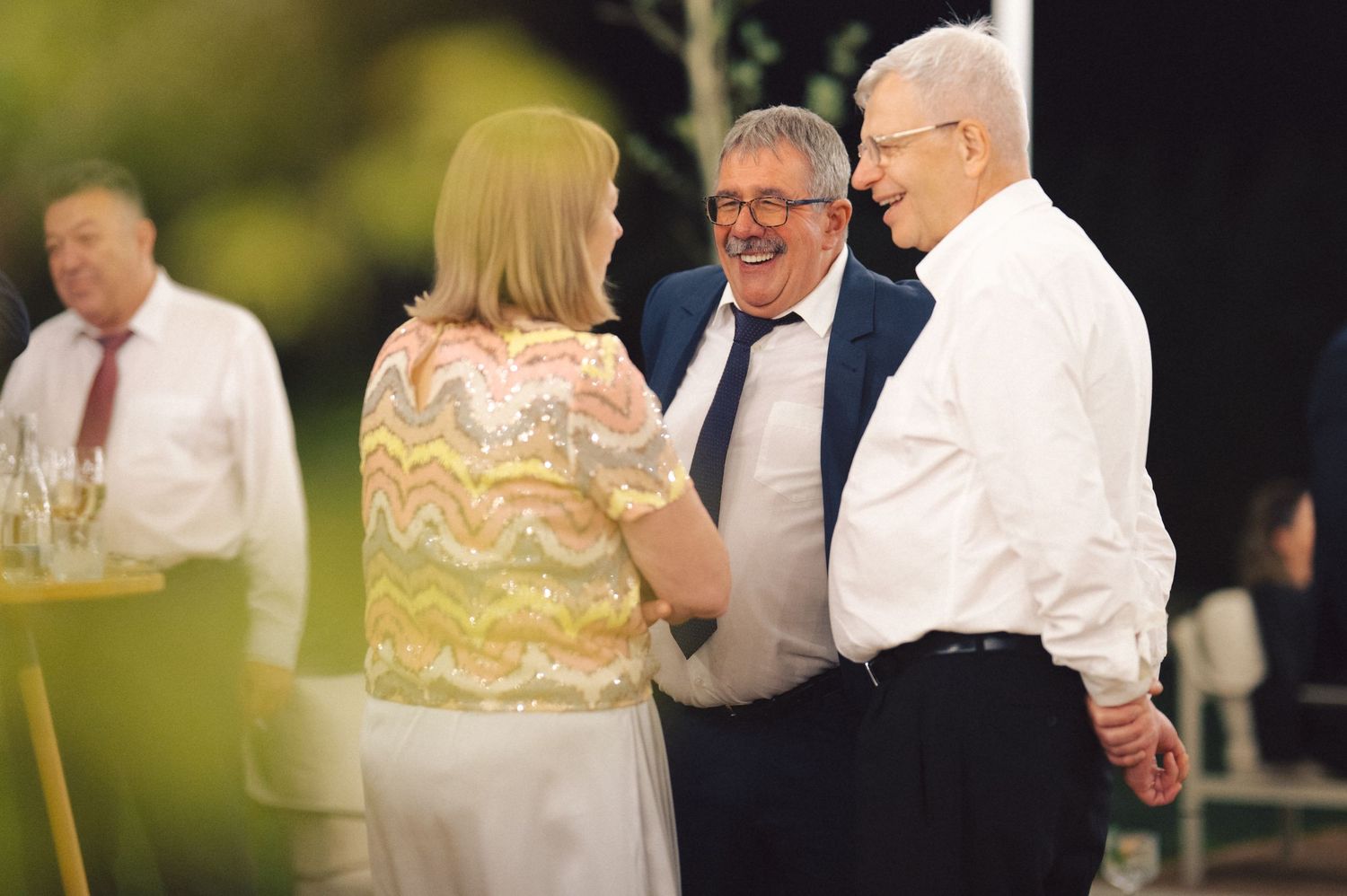 Three people laugh and converse during an outdoor evening celebration.