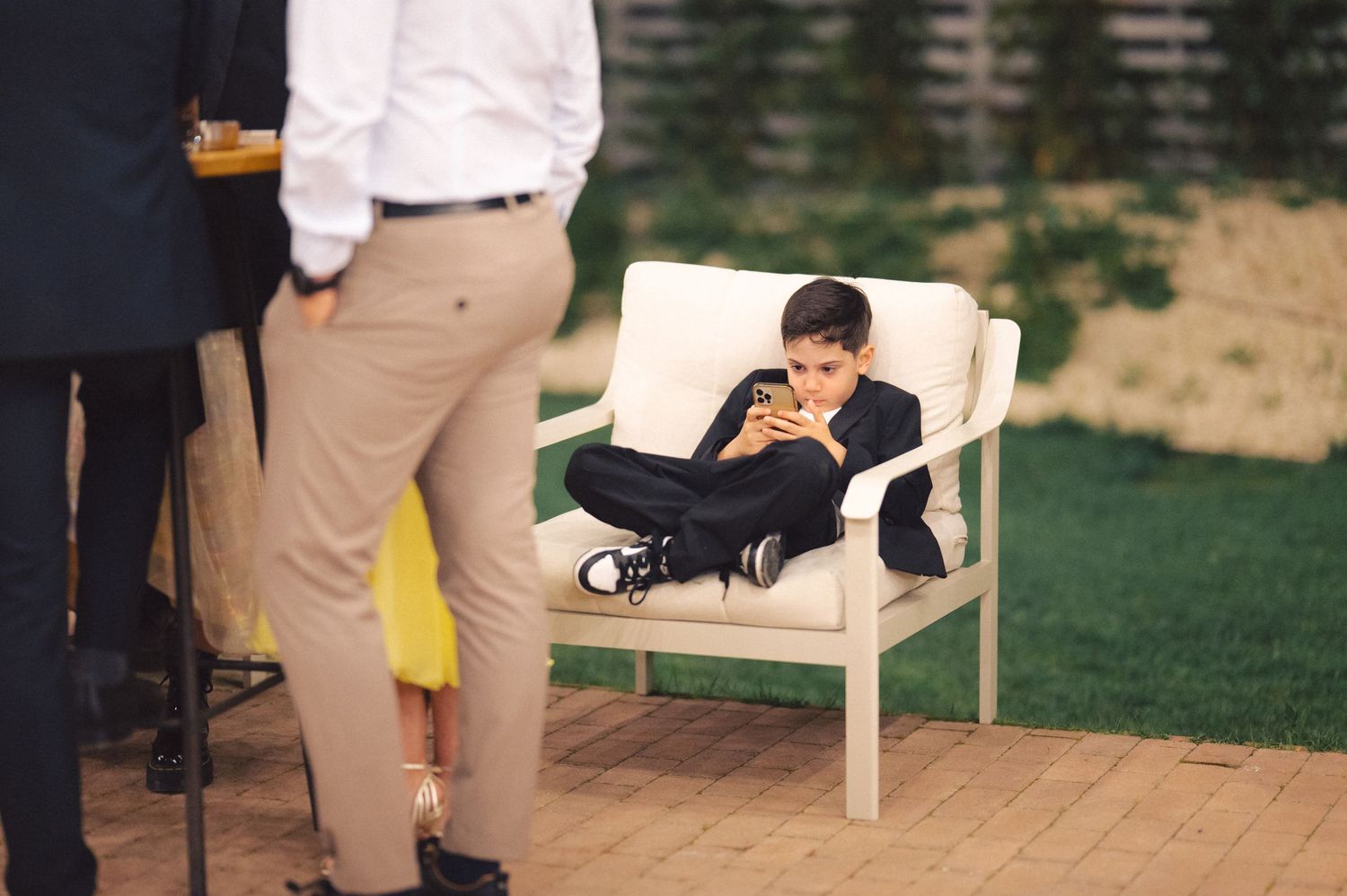 A child reclines on a white outdoor chair on a wooden deck while looking at a mobile device.