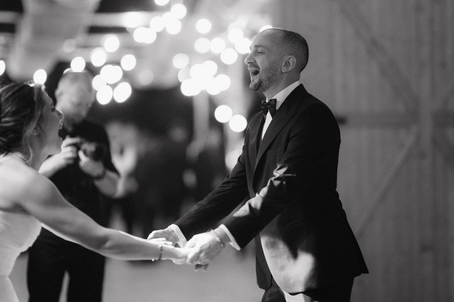Black and white photo of joyful wedding dance moment with tuxedo and lights in background.