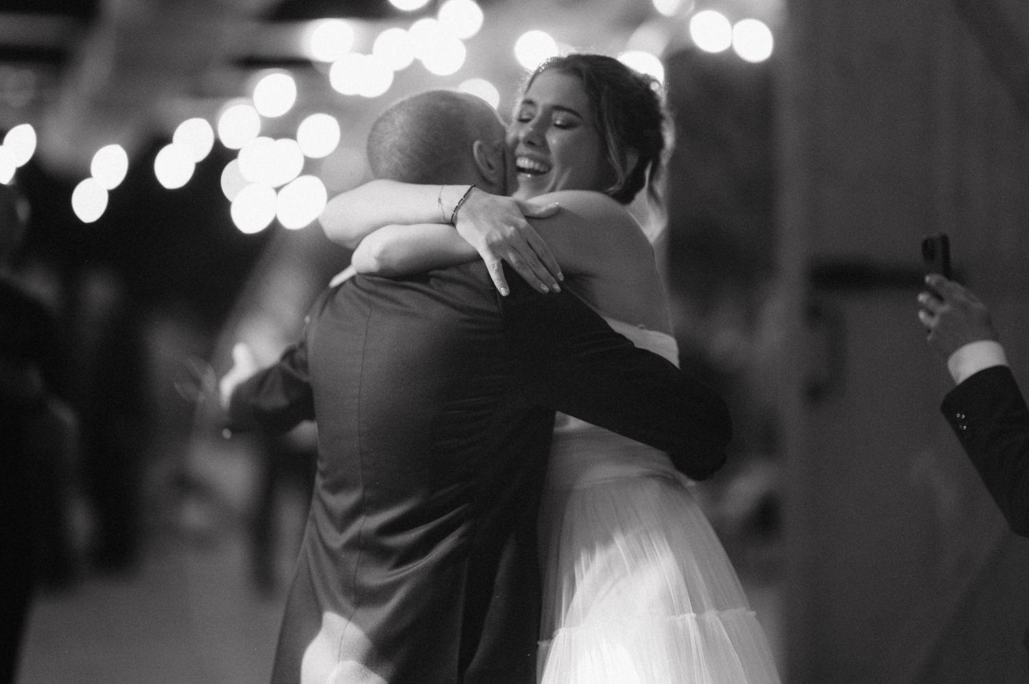 A couple embraces during their first dance at a wedding reception with twinkling lights in the background.