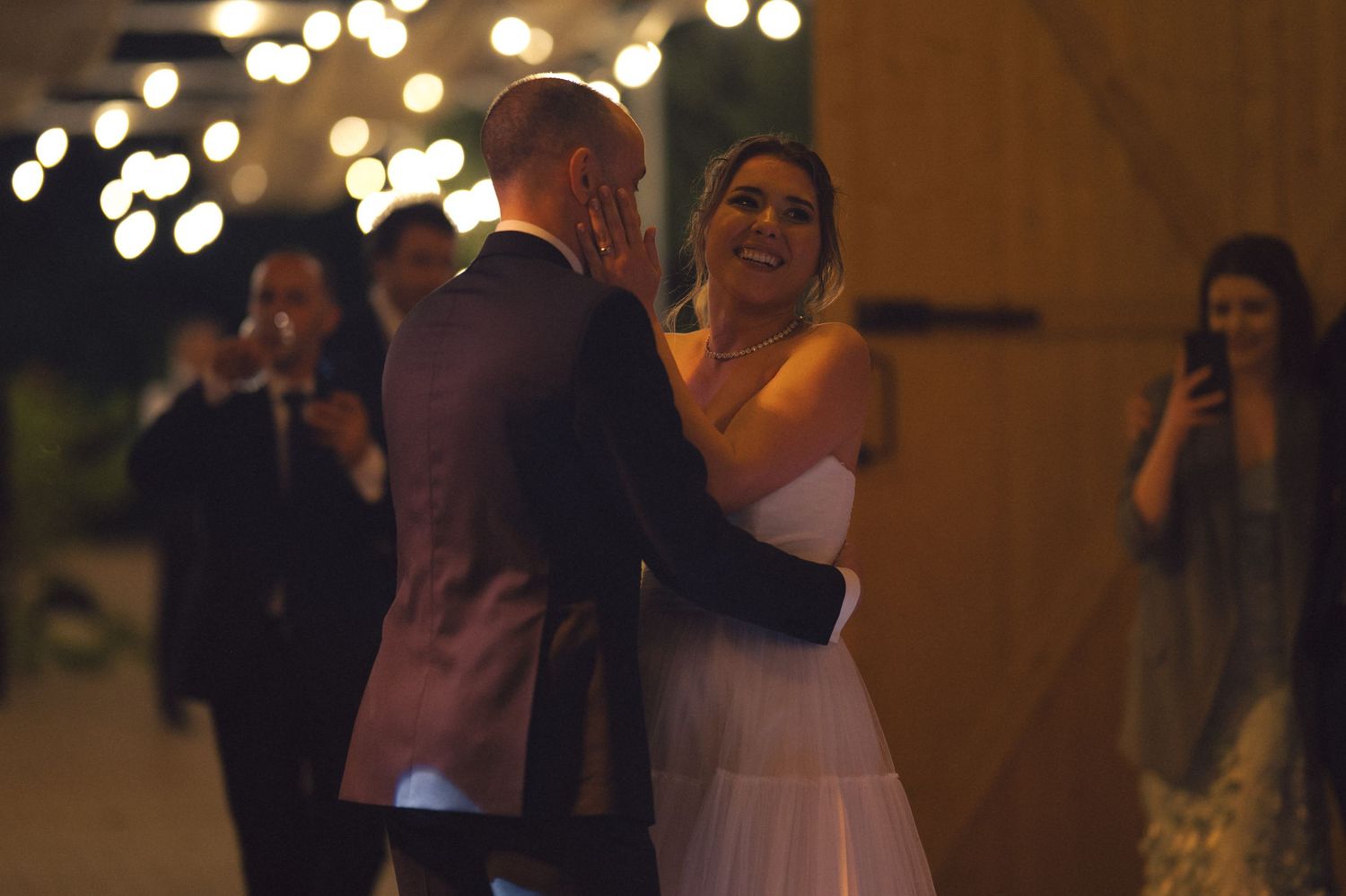 A couple shares a romantic slow dance under glowing lights at their evening wedding reception.