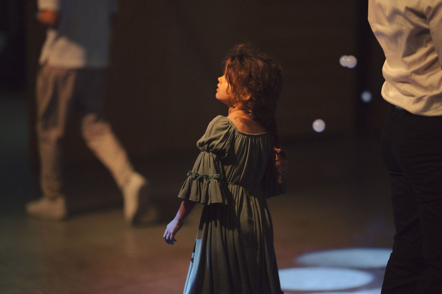 A performer in a flowing green layered dress stands on stage during a theatrical performance.
