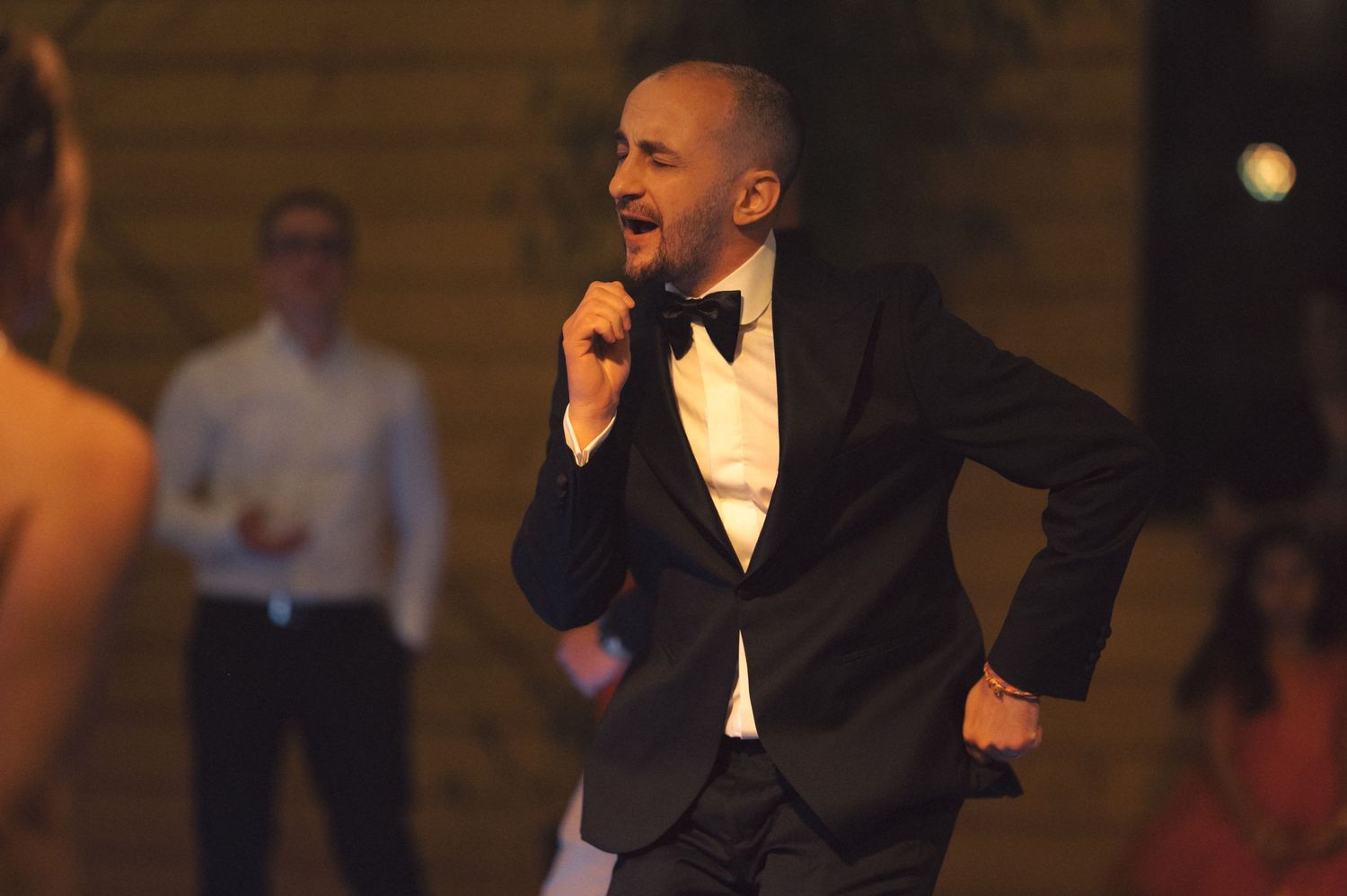 A person in a black tuxedo and bowtie dances expressively at an evening event while others look on in the background.
