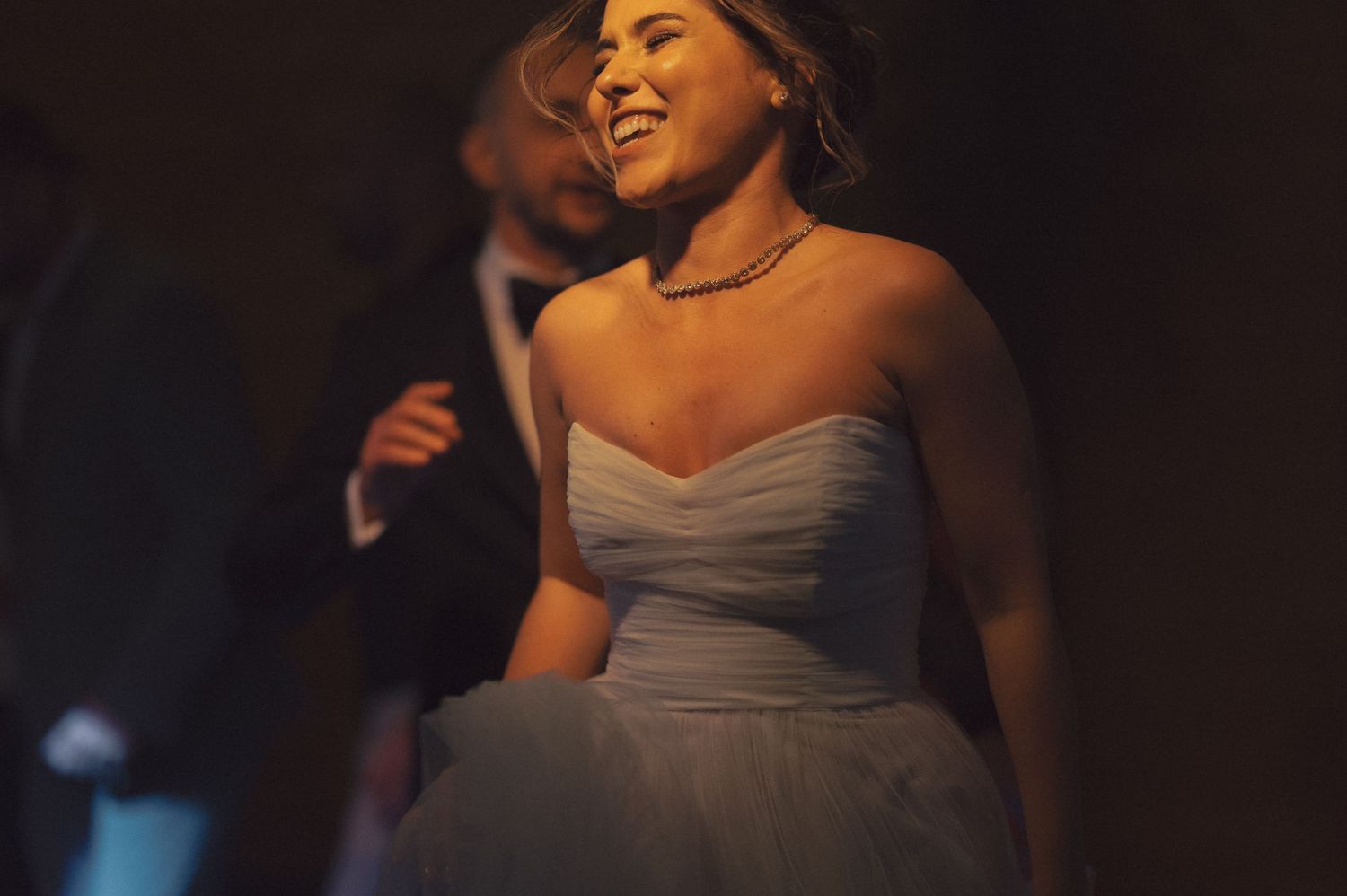 A bride in a strapless white gown laughs joyfully while dancing during an evening wedding reception.
