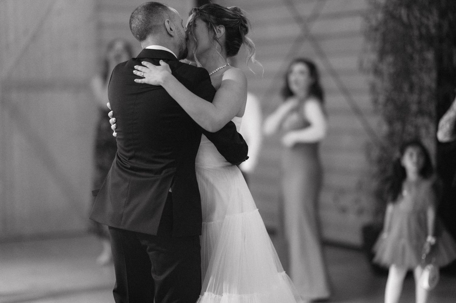 Black and white photo of a romantic first dance at a wedding reception with onlookers in the background.