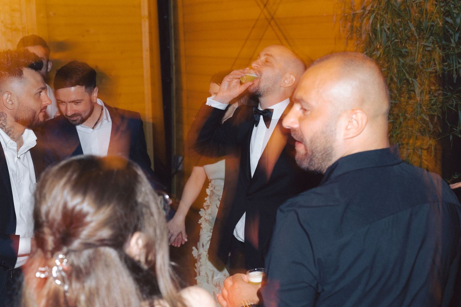 Multiple people standing close together in a dimly lit space with orange lighting while some appear to be drinking.