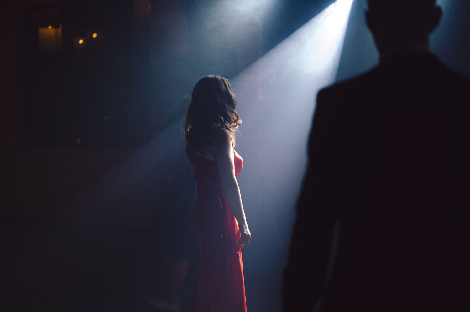 Silhouetted figure stands in dramatic spotlight on dark stage wearing red dress during performance.