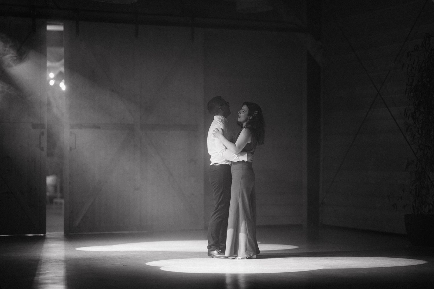 A couple dances intimately in a dramatic spotlight on an empty dance floor, captured in black and white.