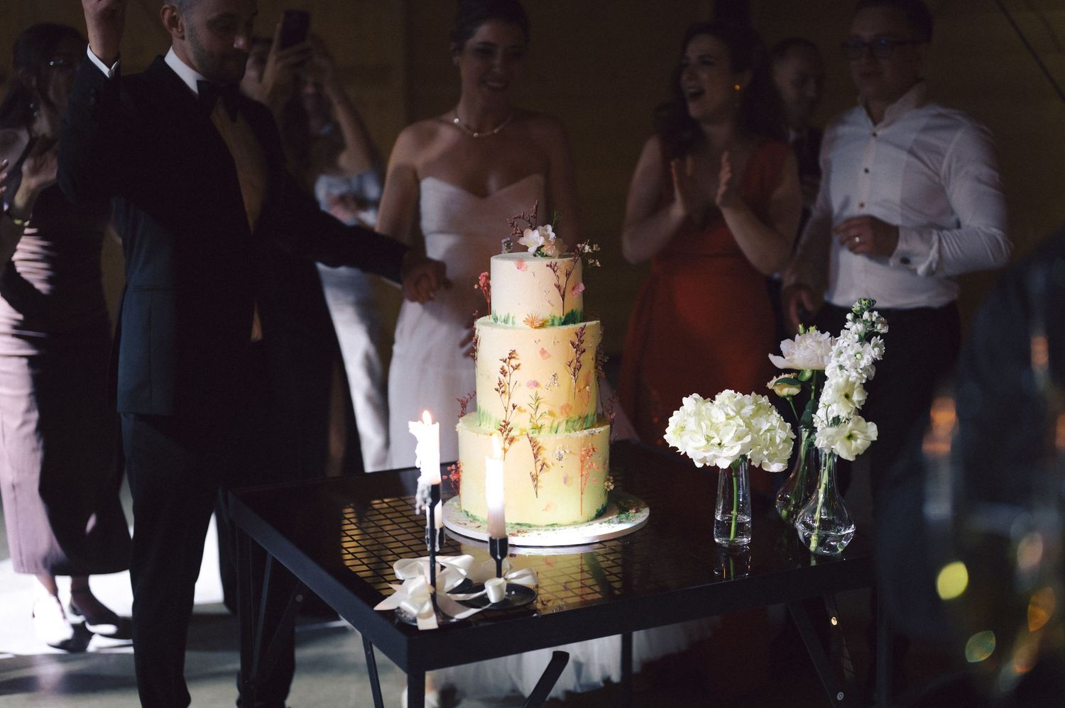 A wedding cake display table with white floral arrangements and guests gathered in a dimly lit celebration setting.