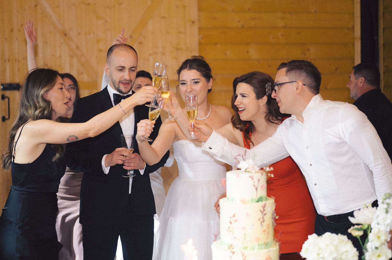 Wedding party guests raise champagne glasses in a toast around a white tiered cake during the reception celebration.