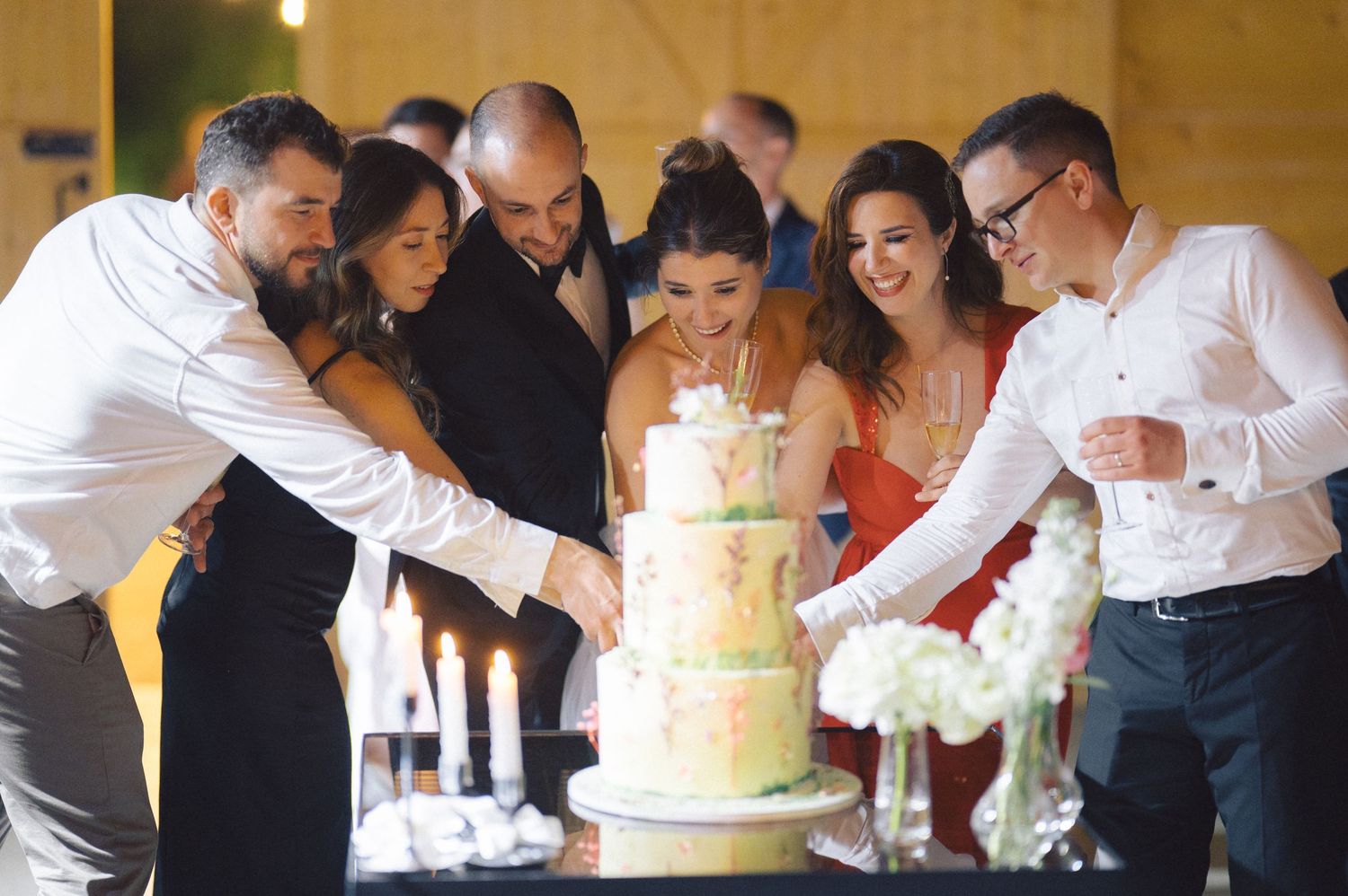 A group of people gather around a tiered wedding cake with lit candles at a celebration.