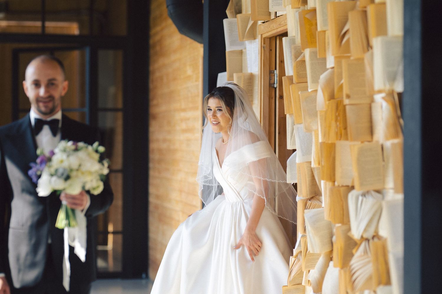 A wedding scene with two figures standing near a wall decorated with hanging cream-colored paper decorations.