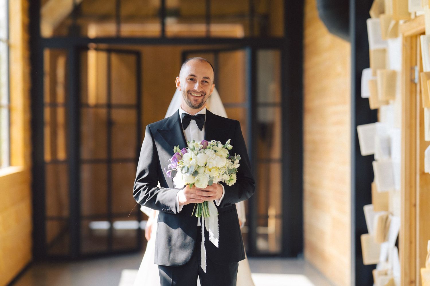 Person in black tuxedo holding white floral bouquet while walking through elegant hallway with arched windows.