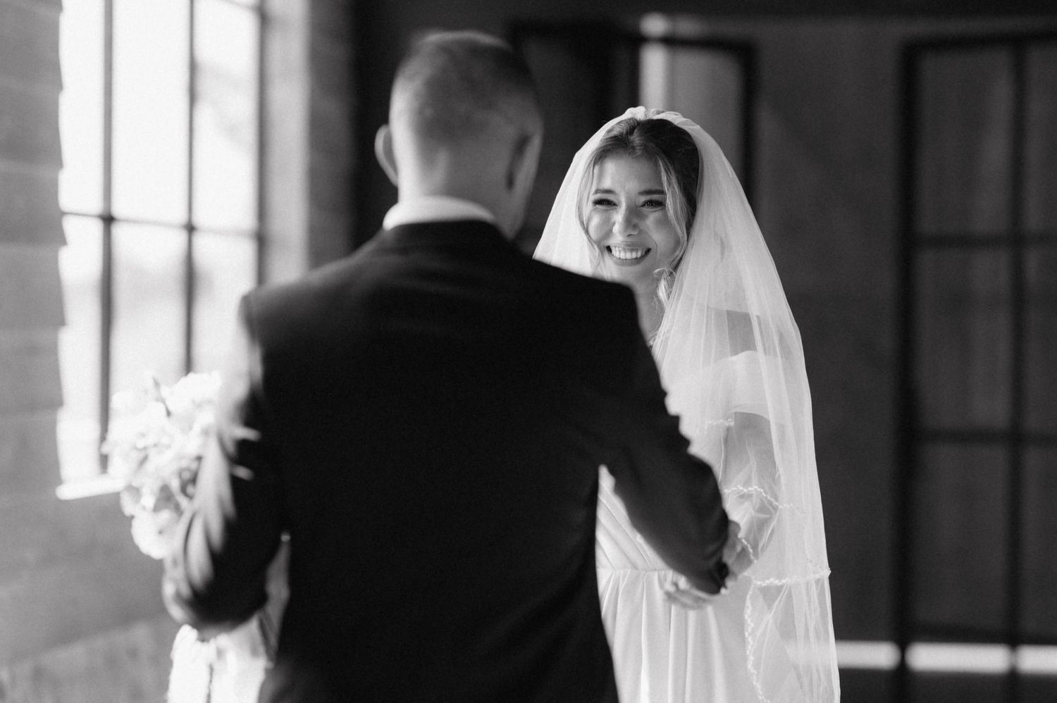 A bride in a white veil smiles over her shoulder while dancing with her partner in a black suit during their wedding.