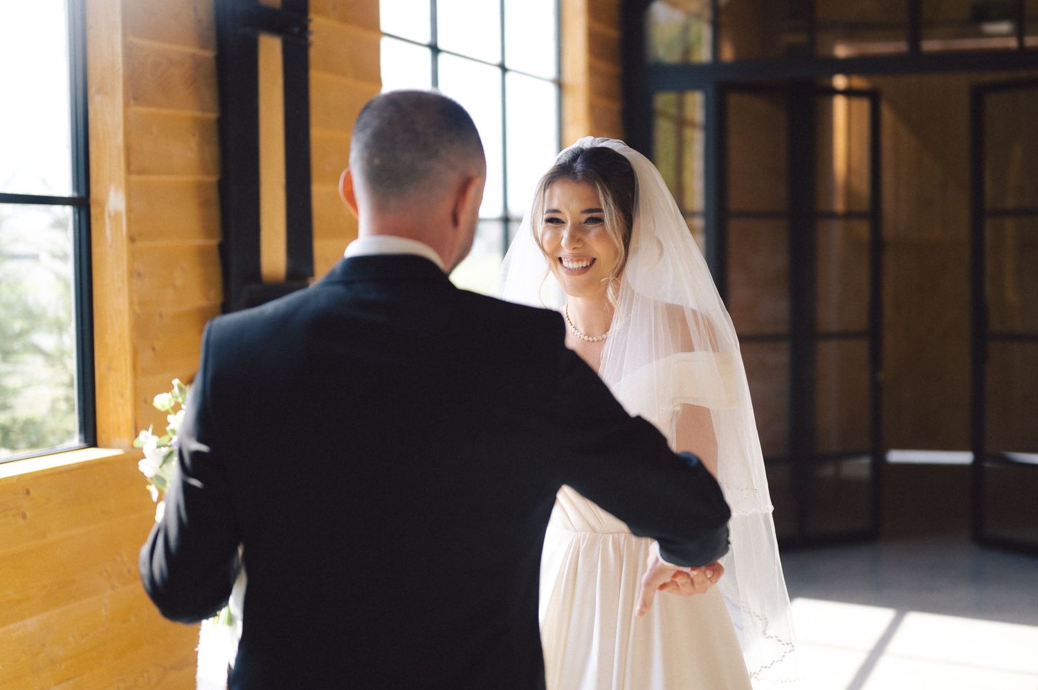 Person in white wedding gown and veil meeting their partner in a sunlit hallway with dramatic windows.