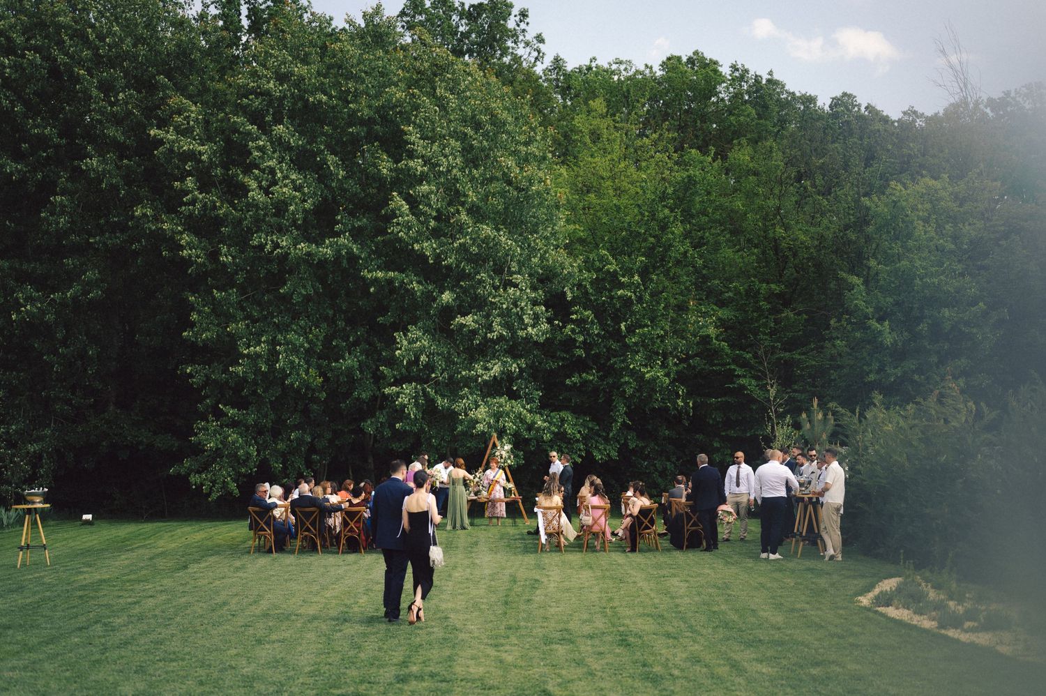 People gather outdoors on a green lawn for a summer event near a wooded area under partly cloudy skies.