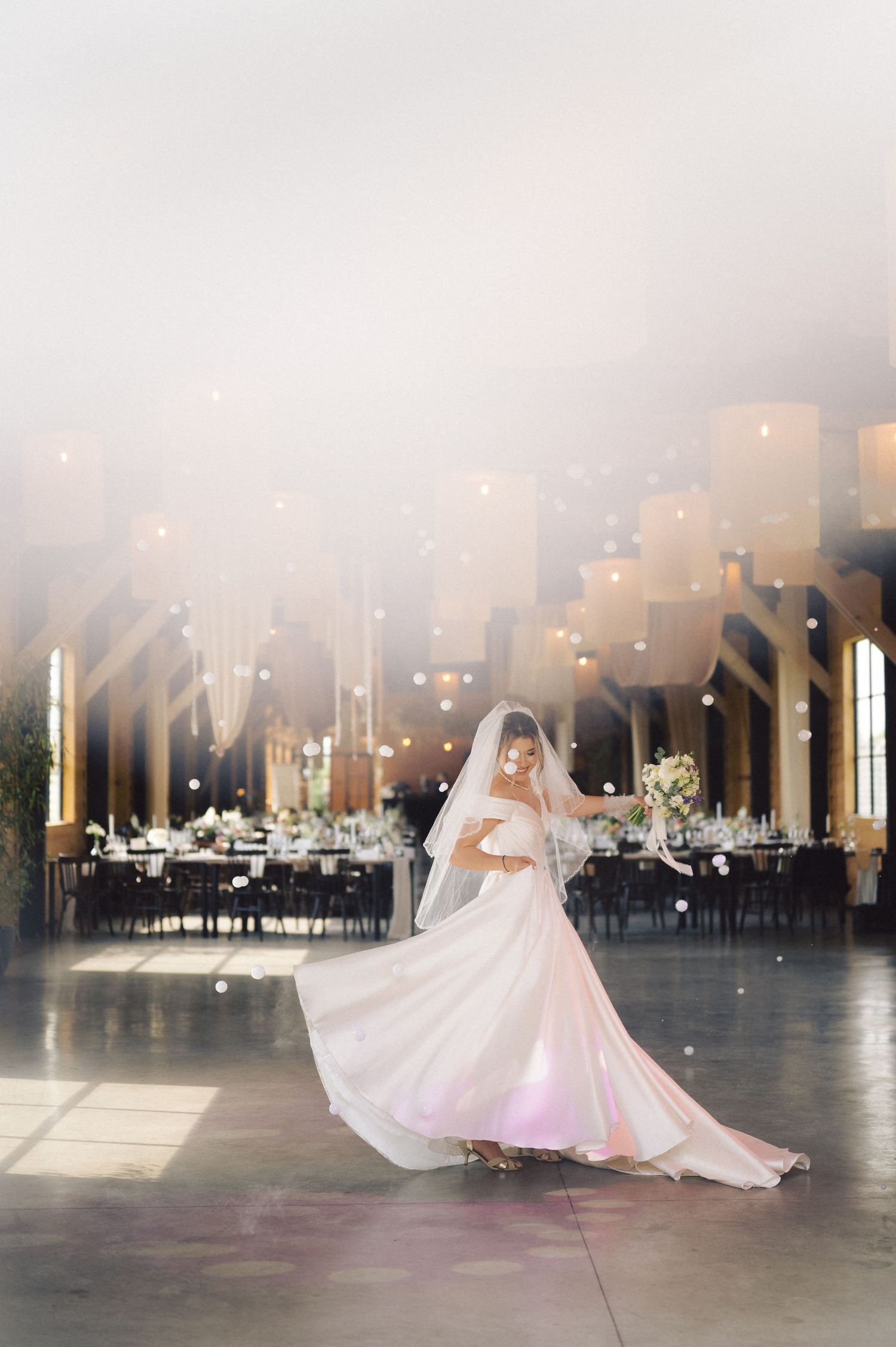 A twirling figure in a flowing white wedding gown and veil stands in an elegant reception venue with glowing lights above.