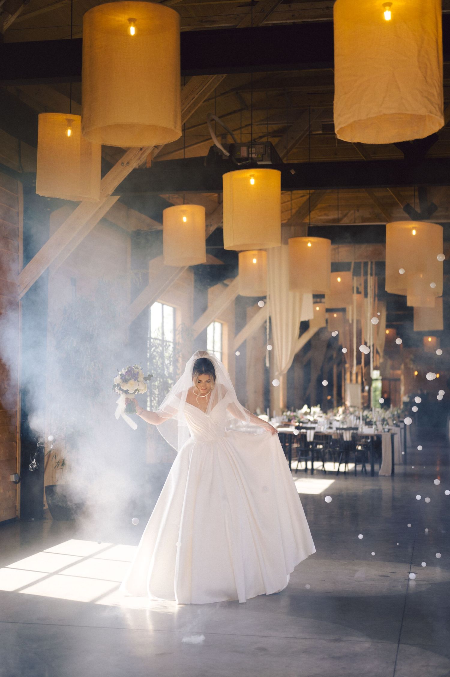 A bride twirls in a white wedding gown under large cylindrical pendant lights in a rustic venue filled with ethereal smoke.