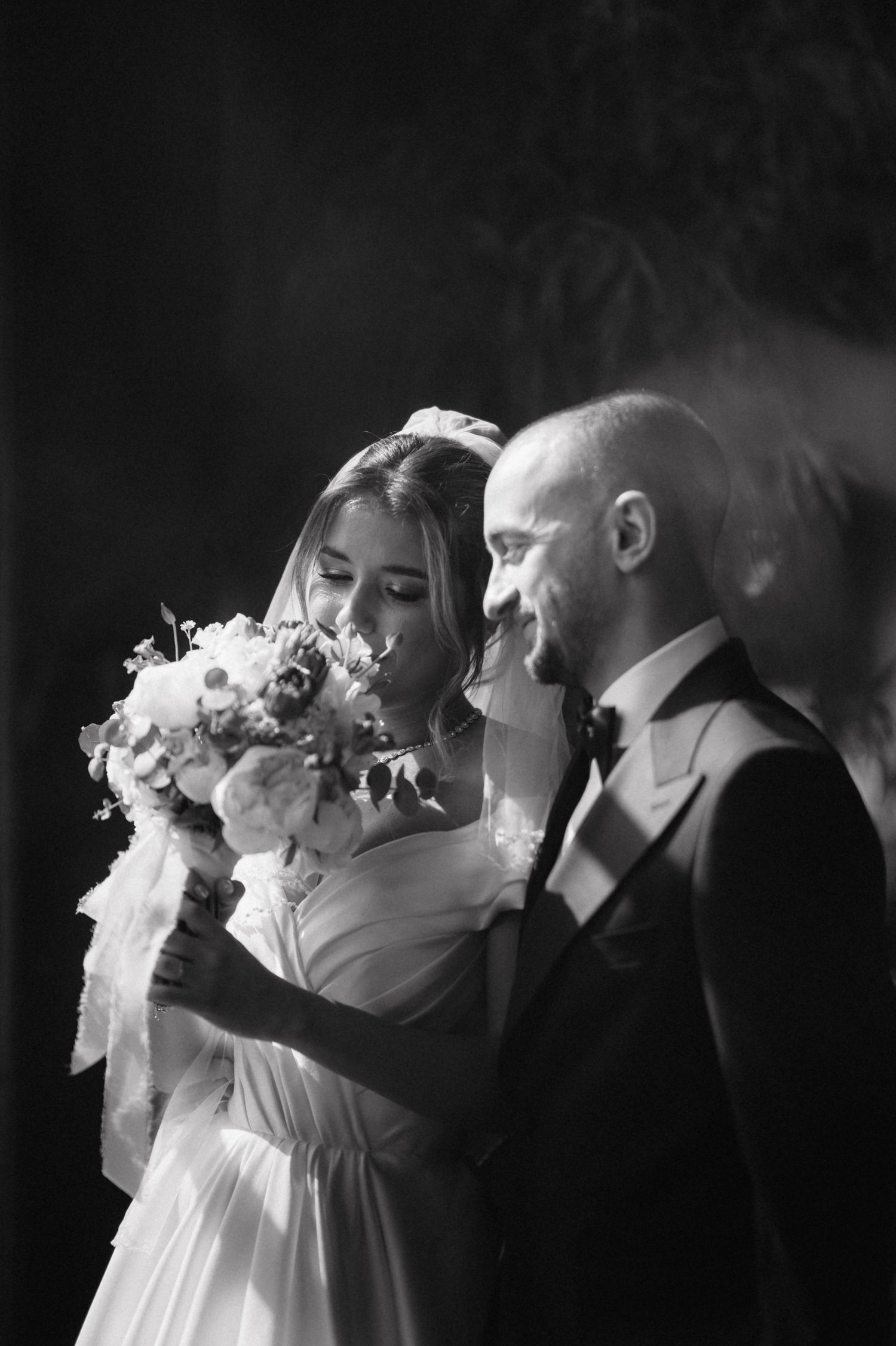 A black and white wedding photograph showing a bride in white dress holding a floral bouquet next to a groom in suit.