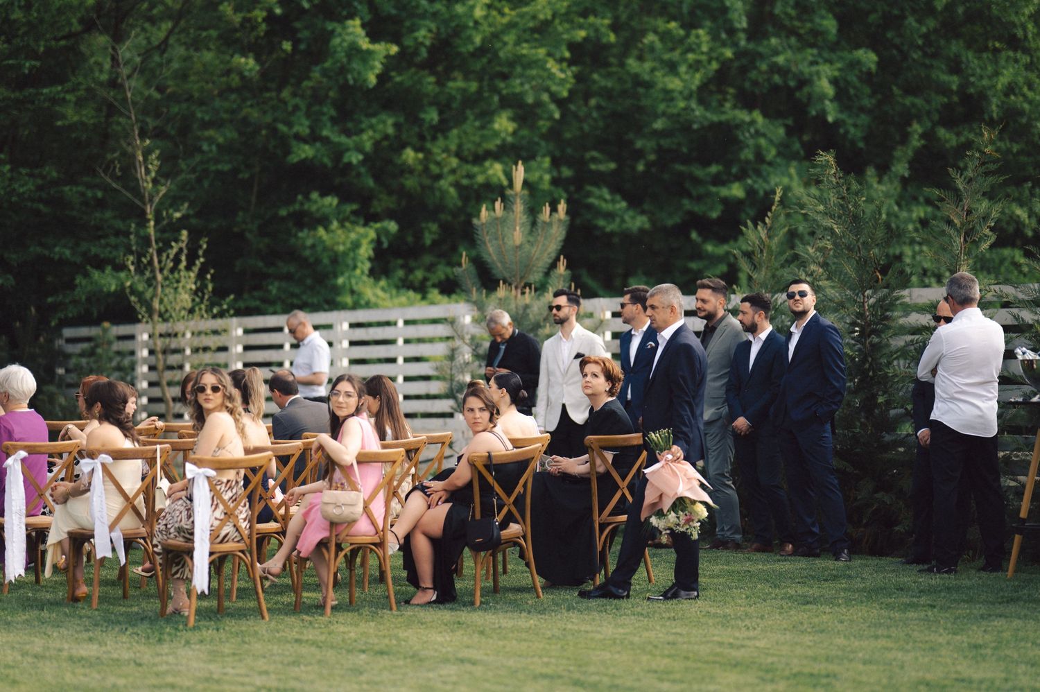 Wedding guests gather on a lush green lawn with white fencing and trees in the background during an outdoor ceremony.