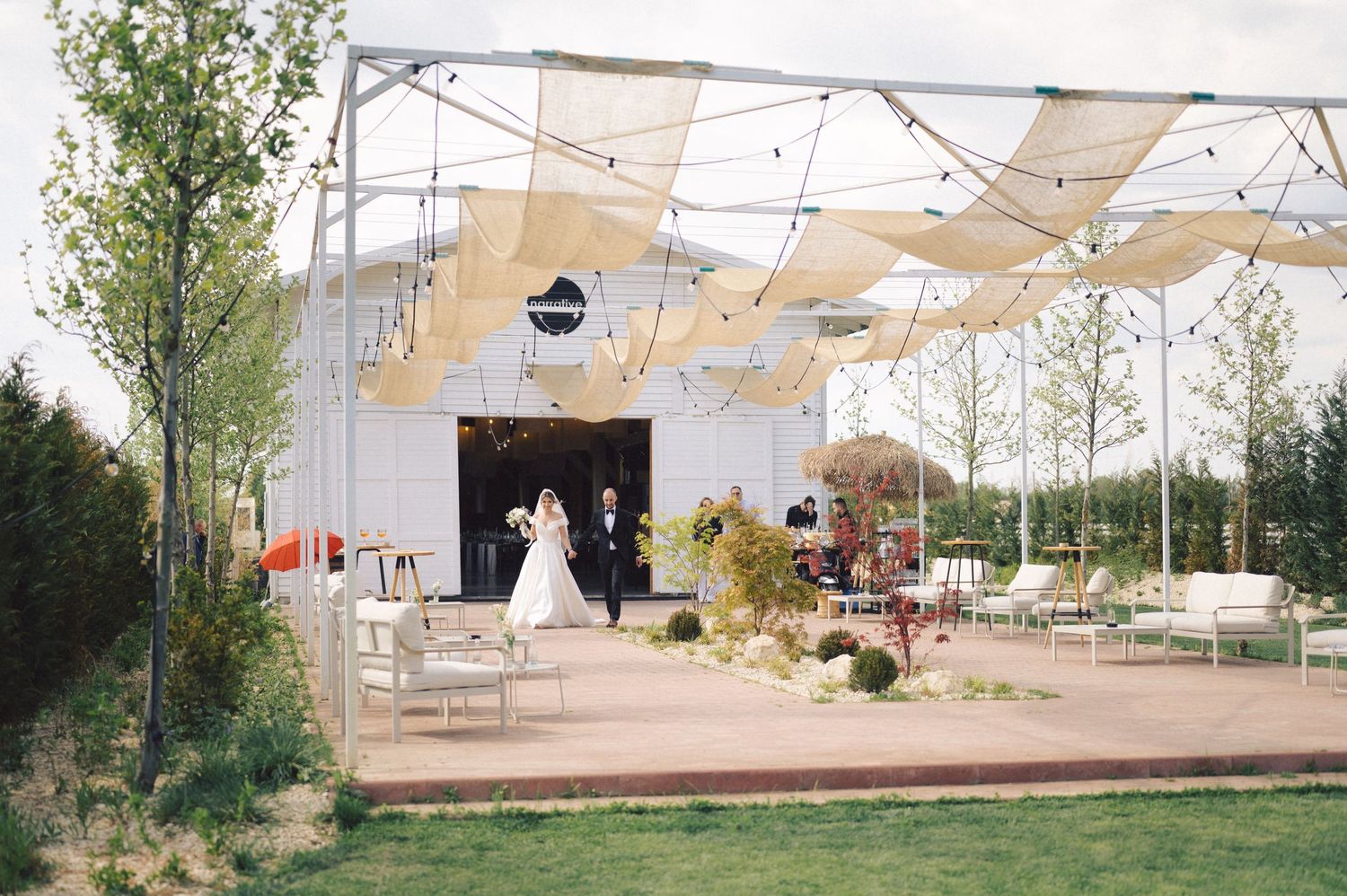 Outdoor wedding venue with light beige canopy sails and white seating arrangements surrounded by trees and greenery.