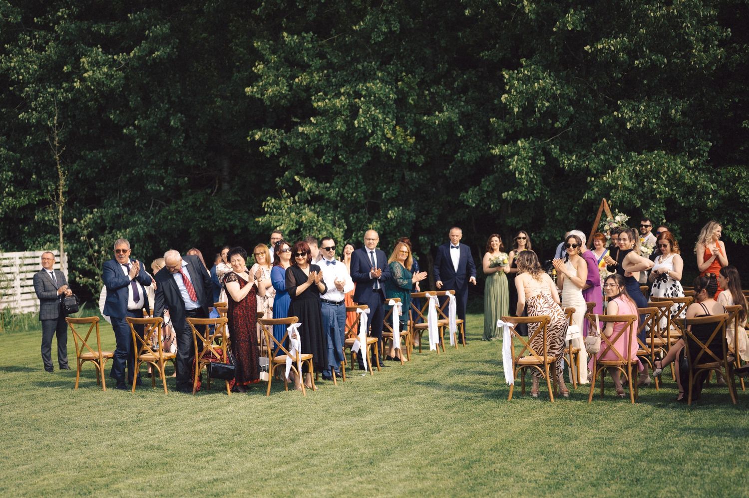 Wedding guests seated on wooden chairs on a lush green lawn during an outdoor summer ceremony.