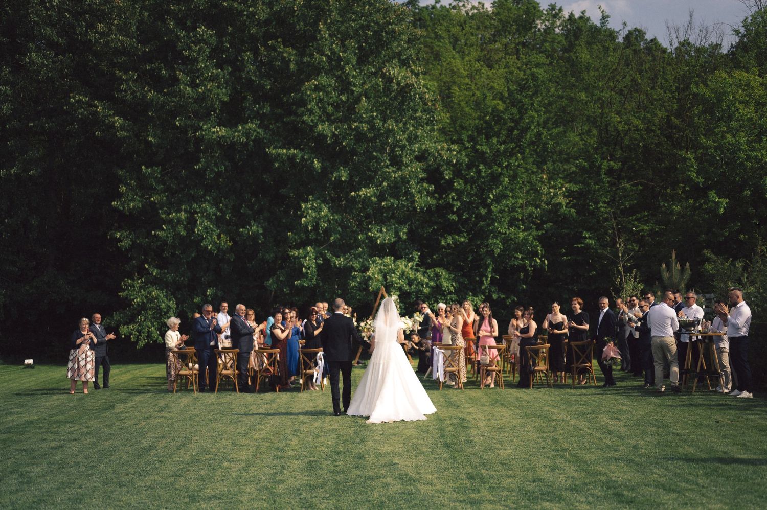 An outdoor wedding ceremony takes place on a lush green lawn surrounded by tall trees and guests gathered in attendance.