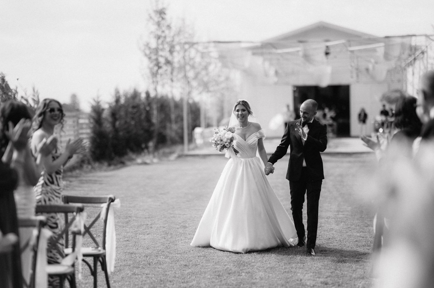 A newlywed couple walking down an outdoor aisle at their wedding venue while guests look on in black and white photo.