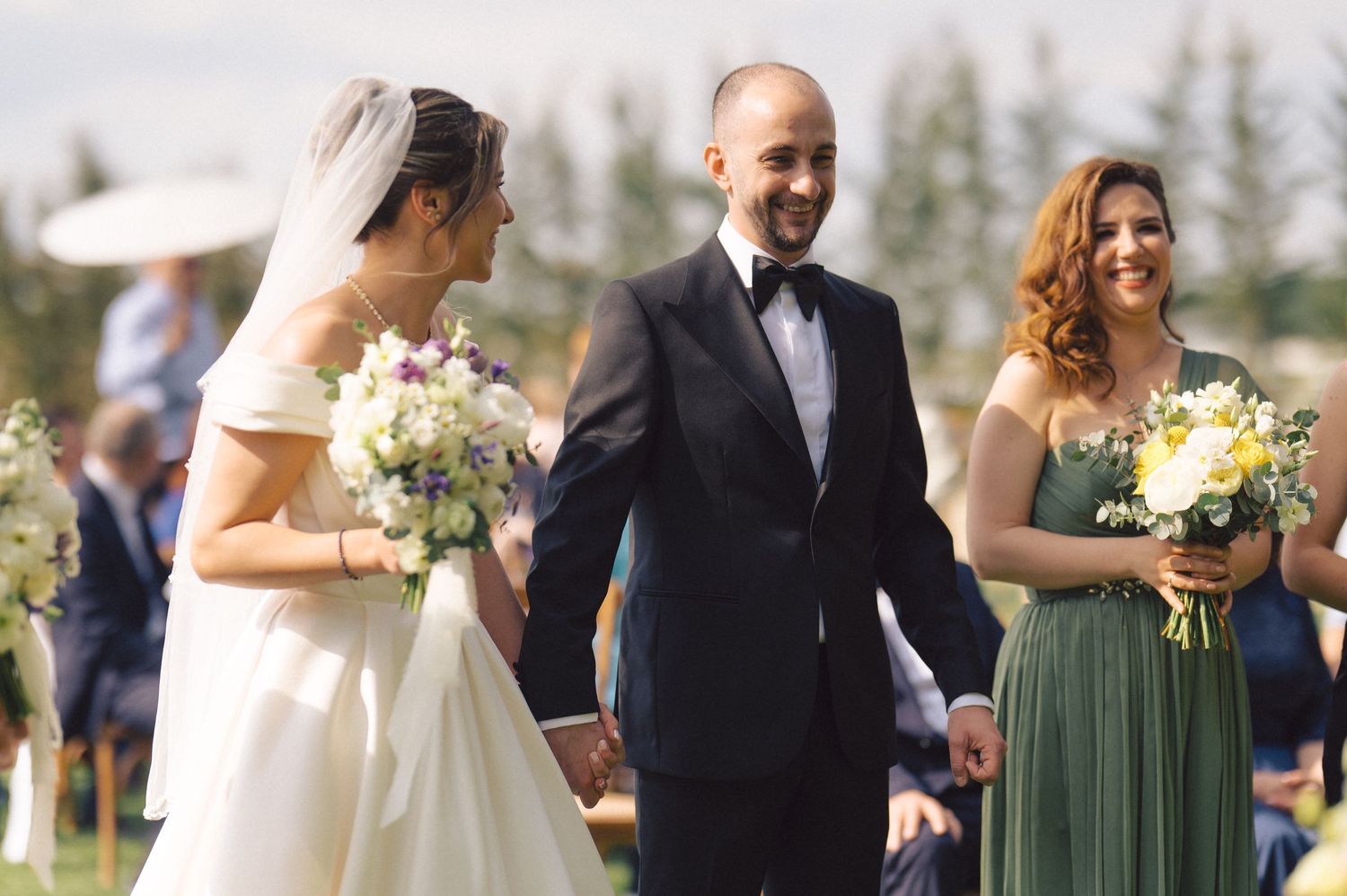 An outdoor wedding ceremony with a bride in white, bridesmaid in green, and groom in black tuxedo holding bouquets.
