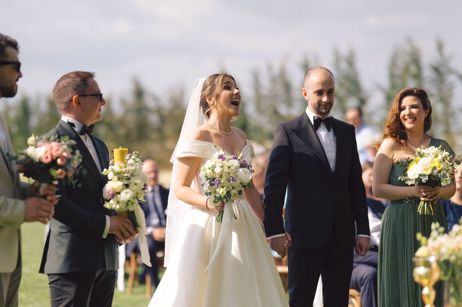 Wedding party stands outdoors holding floral bouquets against a backdrop of evergreen trees on a sunny day.