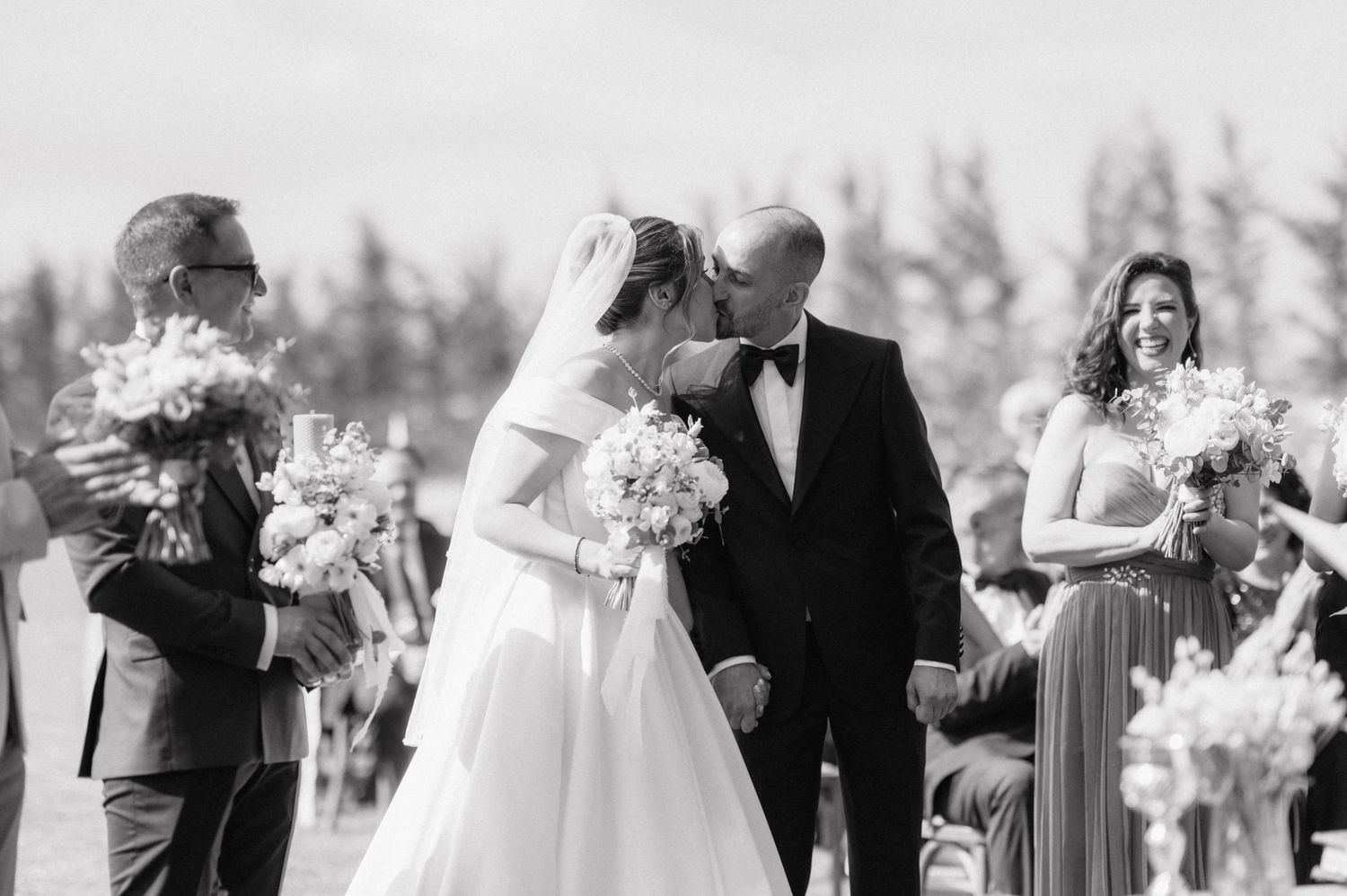 A black and white outdoor wedding ceremony moment captured in front of trees.