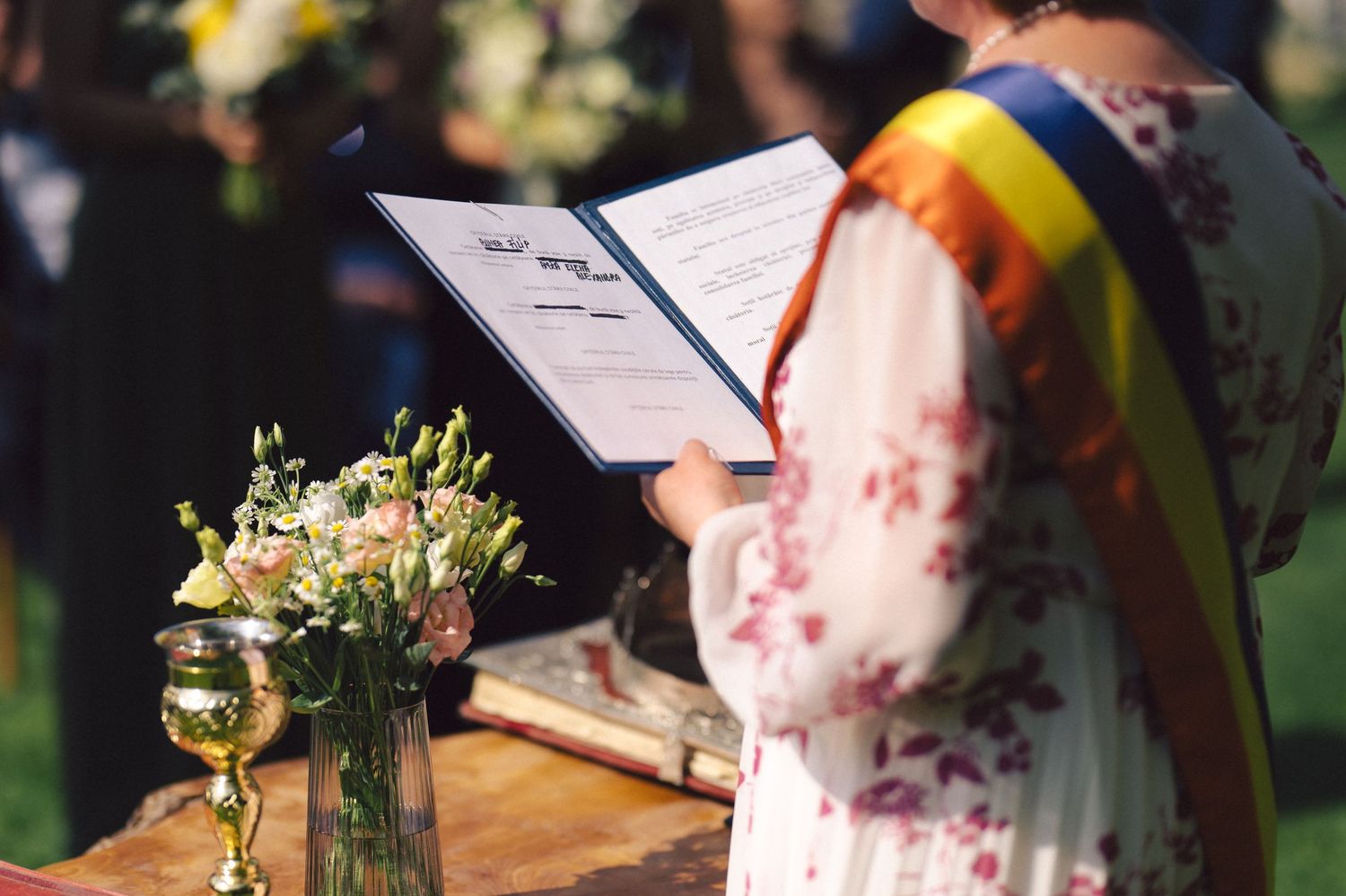 A wedding ceremony moment with a floral centerpiece vase in foreground and a person reading from papers in sunlight.