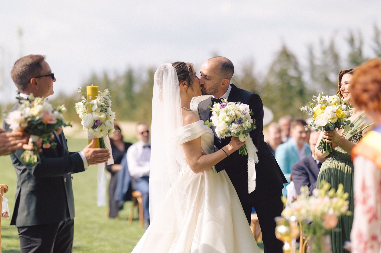 A romantic wedding ceremony outdoors with white and yellow floral bouquets in a sunny garden setting.