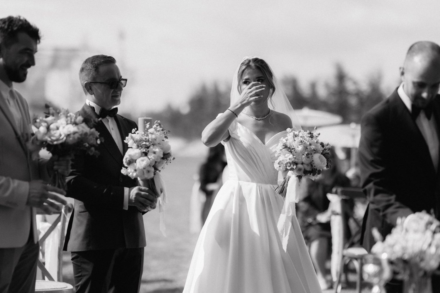 Black and white photo from a wedding ceremony showing an emotional moment with figures holding floral bouquets outdoors.