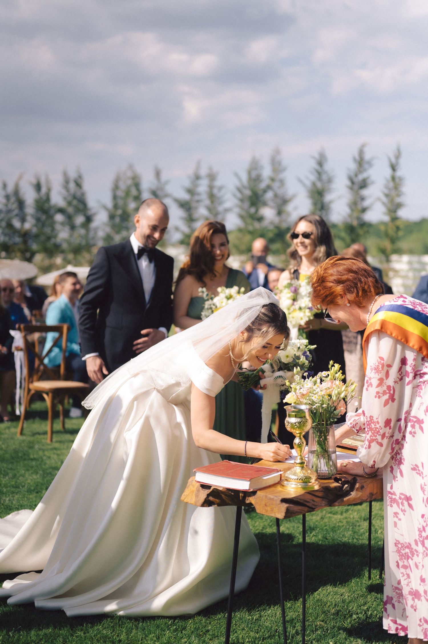 A beautiful outdoor wedding ceremony with a bride in a flowing white gown signing documents on a rustic wooden table.