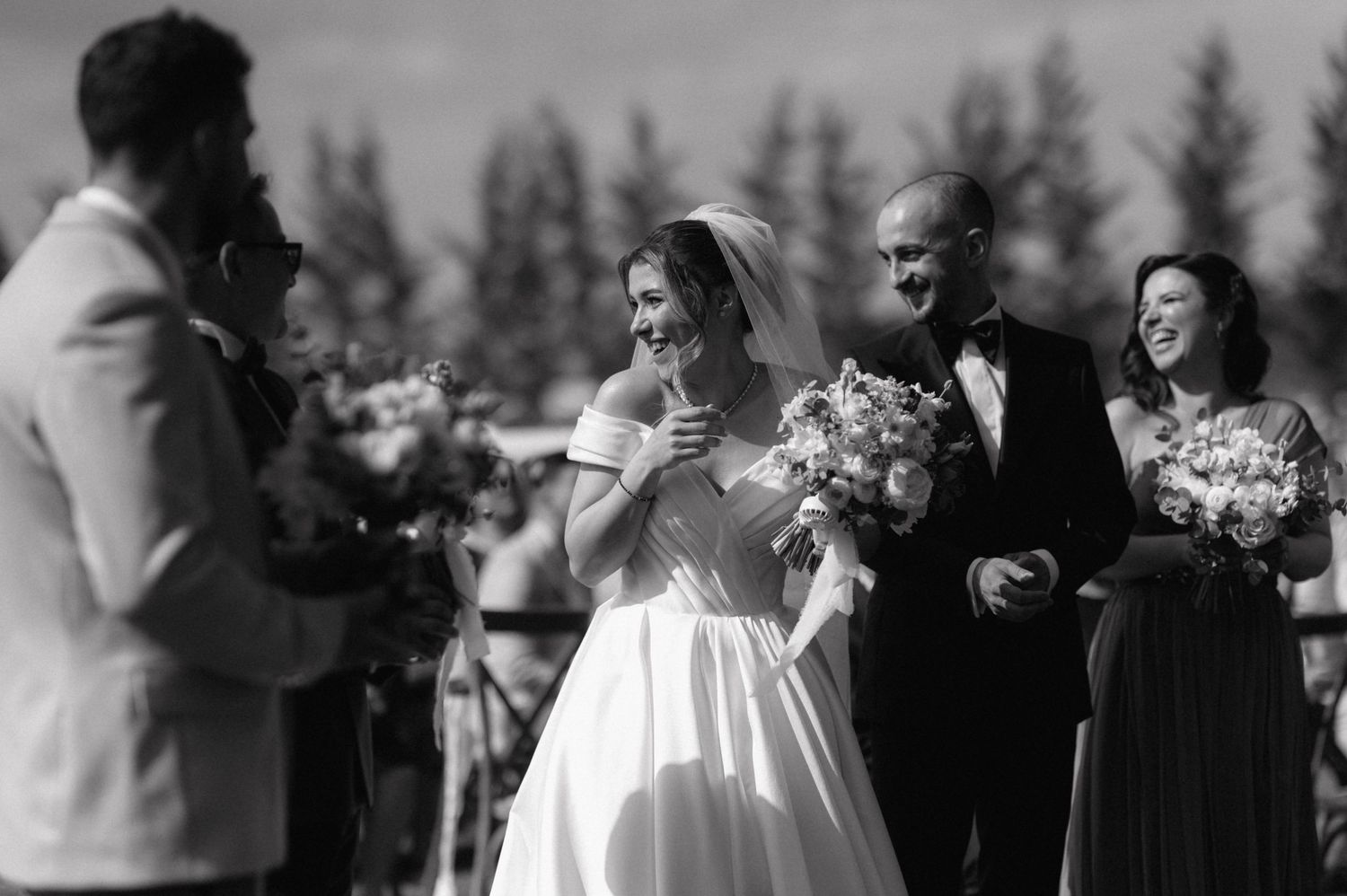 A group of elegantly dressed wedding attendees share joyful moments during an outdoor ceremony.