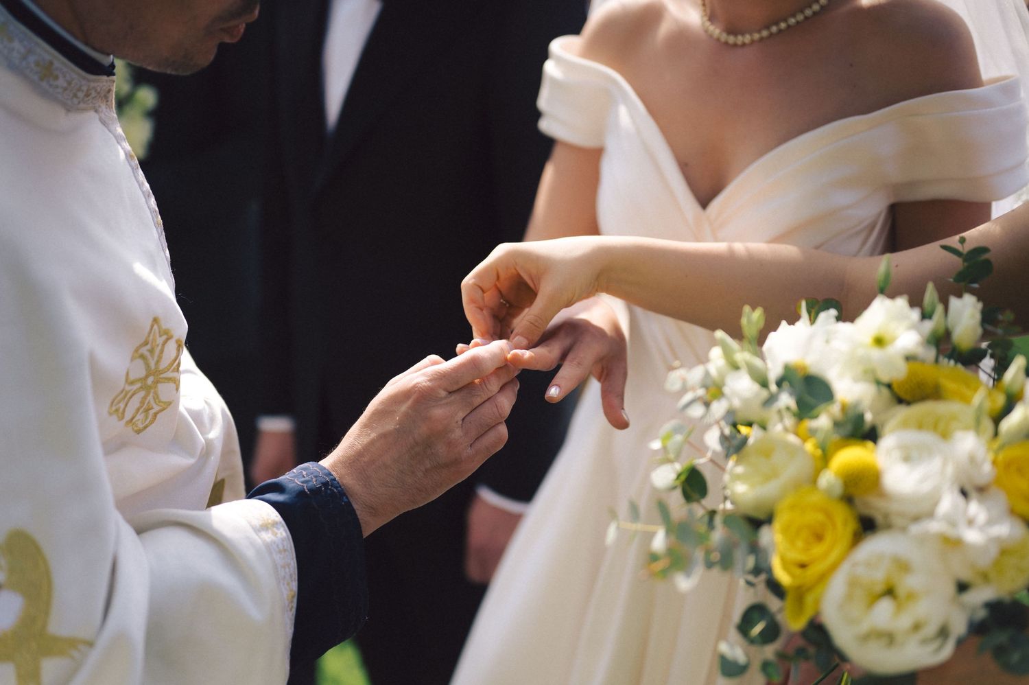 Close-up view of wedding ceremony with white bridal dress, yellow and white bouquet, and exchanging of rings.