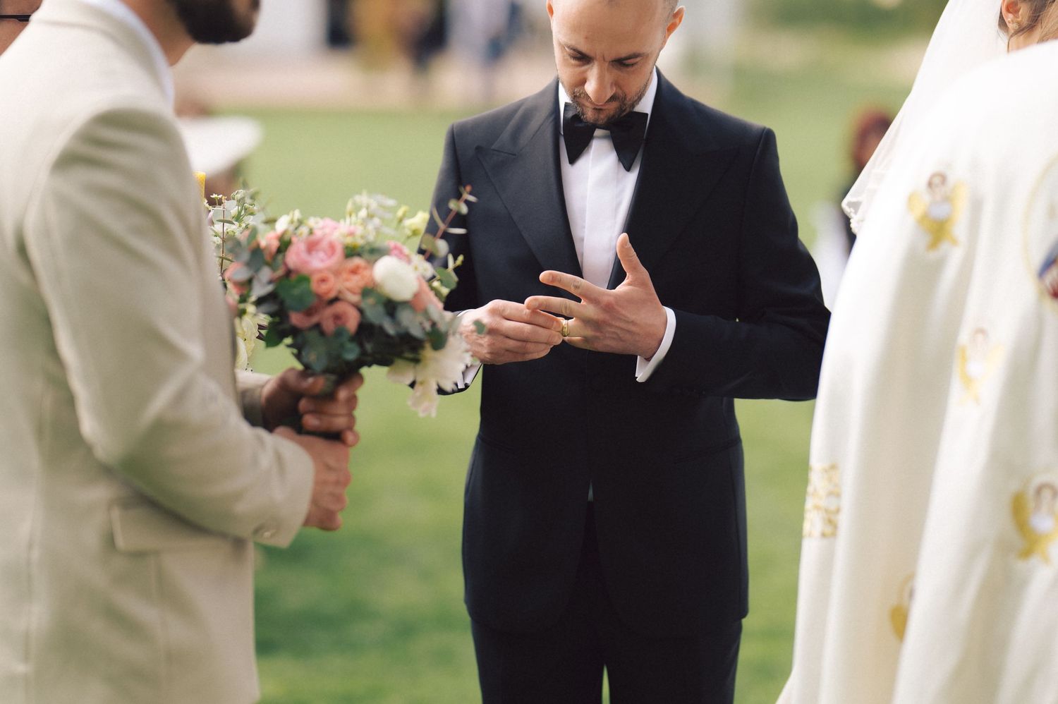 A wedding party member in a black tuxedo checks his phone while standing outdoors during the ceremony.