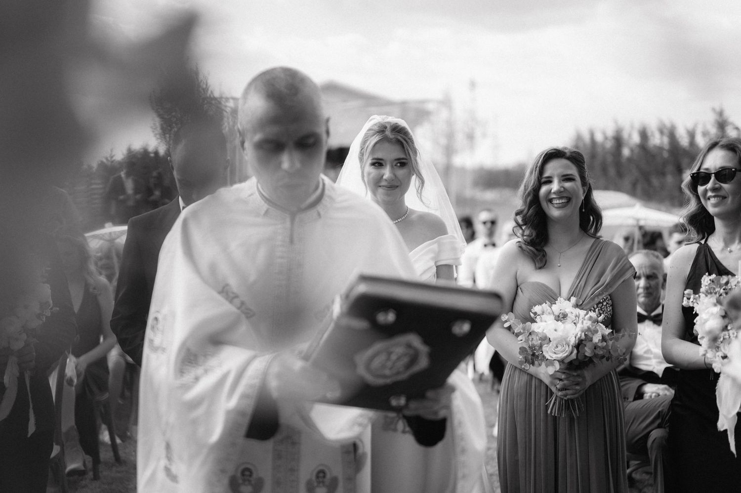 A black and white wedding ceremony photo showing guests and wedding party members gathered outdoors in bright sunlight.