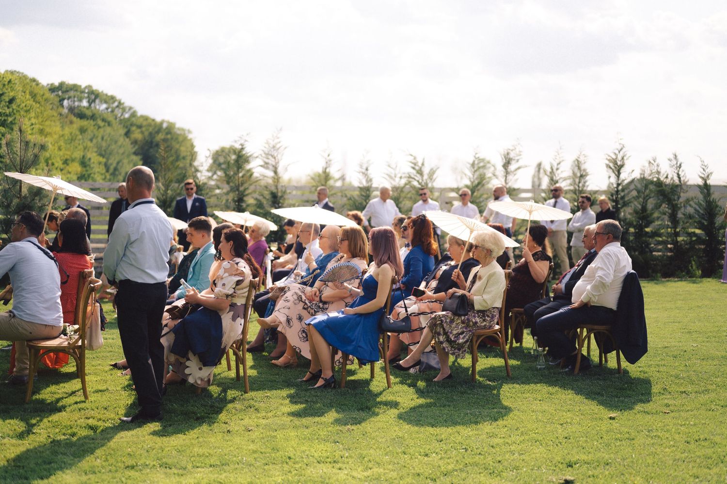 Wedding guests seated on white chairs on a sunny lawn during an outdoor ceremony.