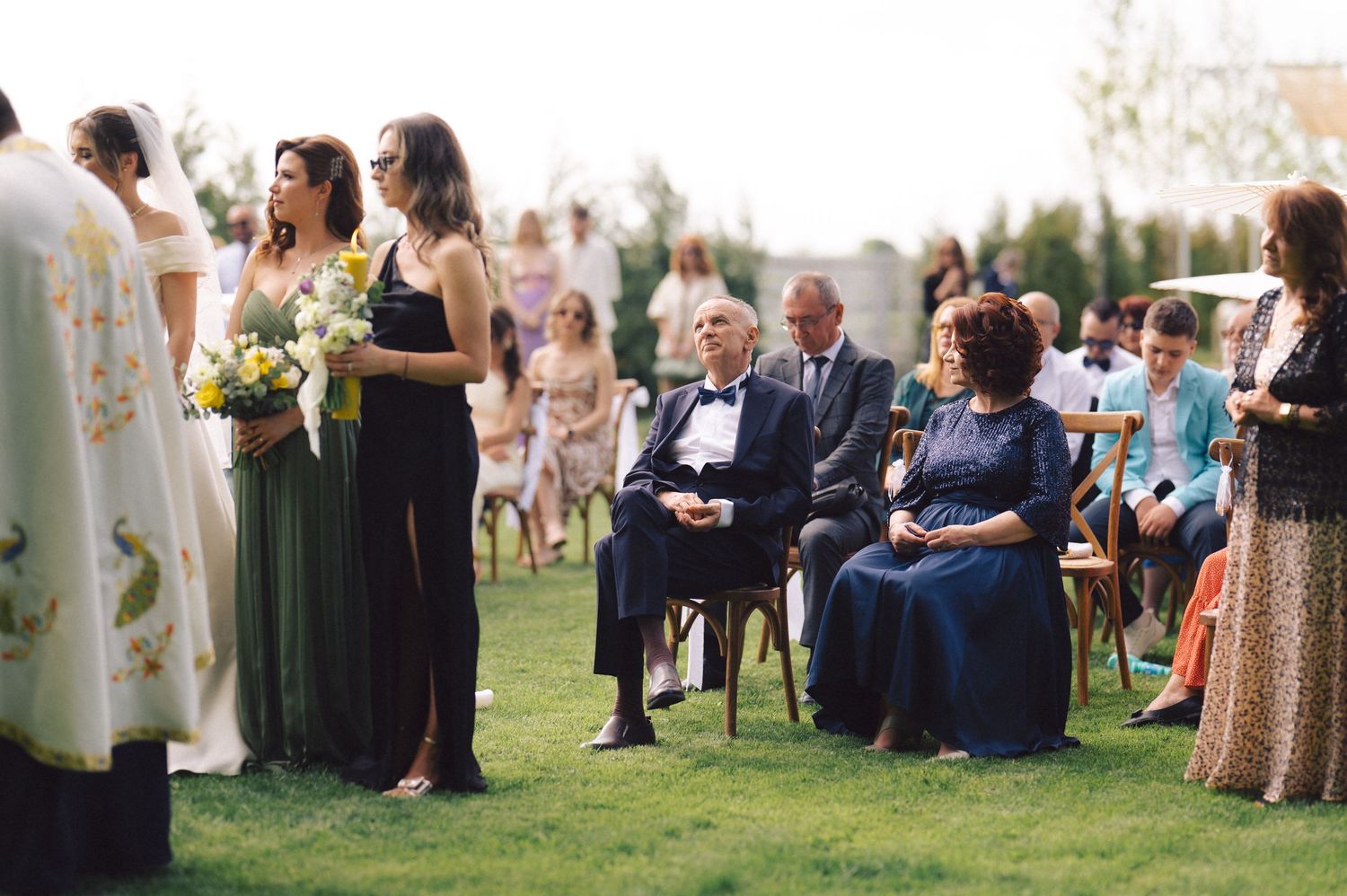 Wedding guests gather on a lawn during an outdoor ceremony, with people seated and standing in formal attire at sunset.