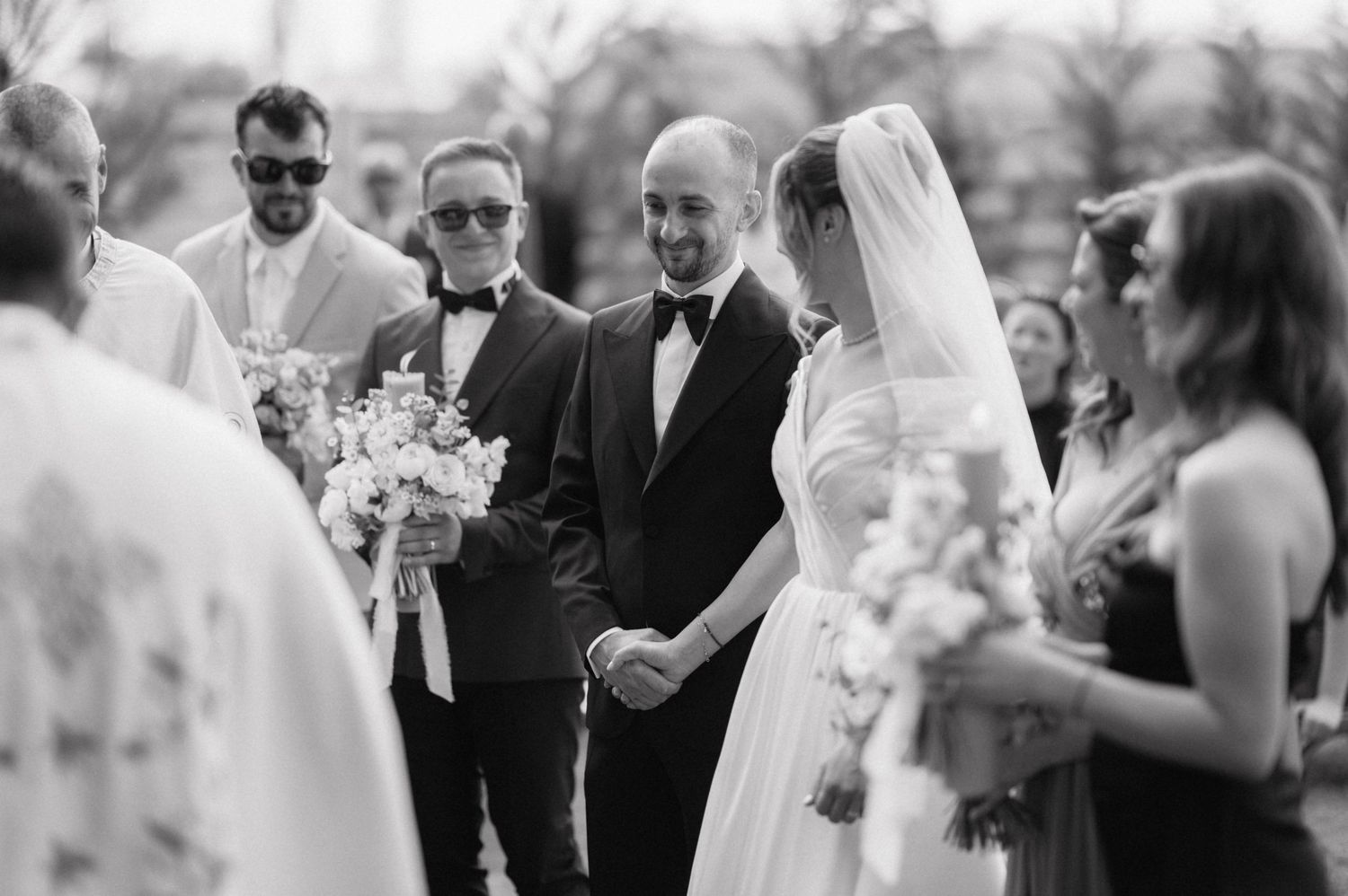 A black and white photograph of an outdoor wedding ceremony with guests gathered around the bride and groom.
