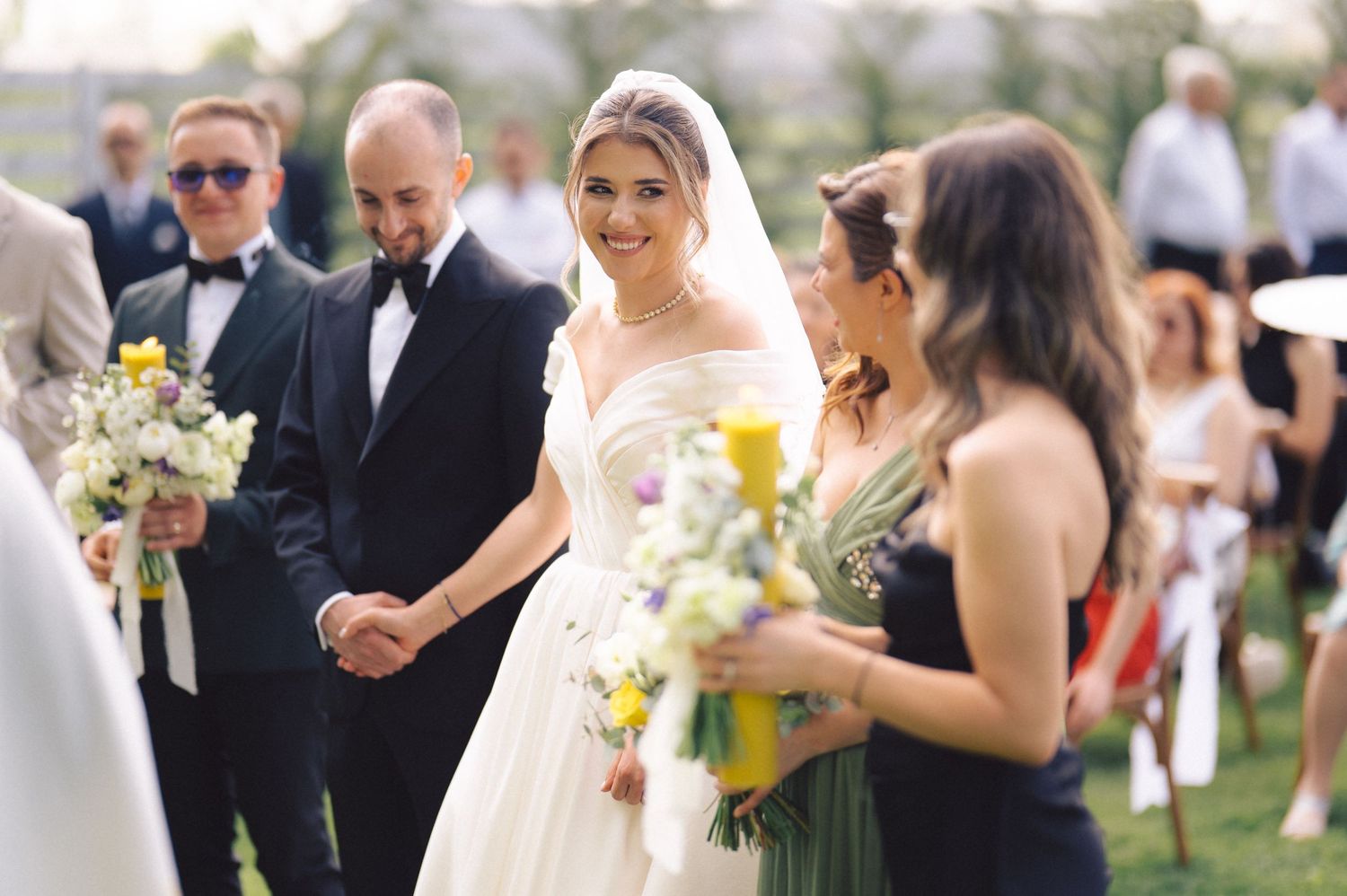 Wedding party members stand together outdoors holding bouquets during a sunny afternoon celebration.