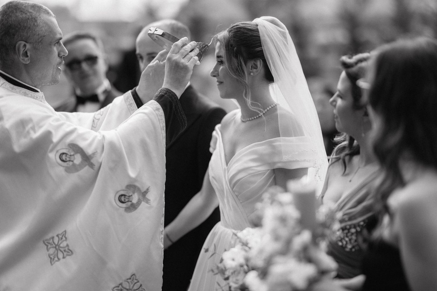 A black and white religious ceremony moment showing the bride receiving a blessing from the officiant.