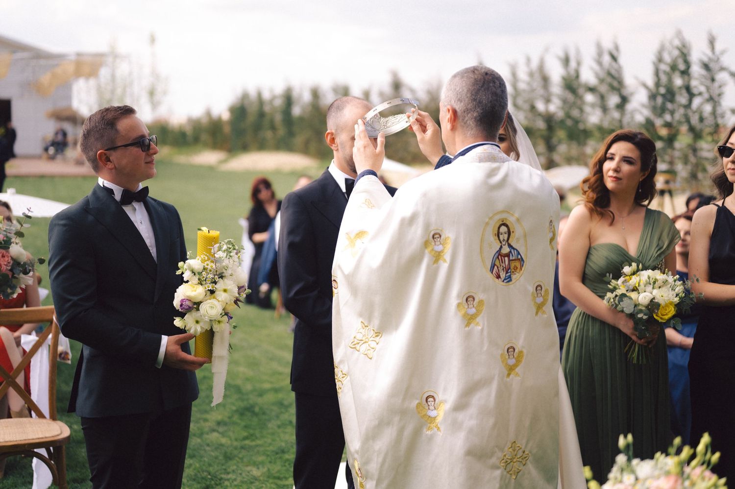 A priest in white robes performs an outdoor wedding ceremony surrounded by guests holding flowers on a grassy lawn.