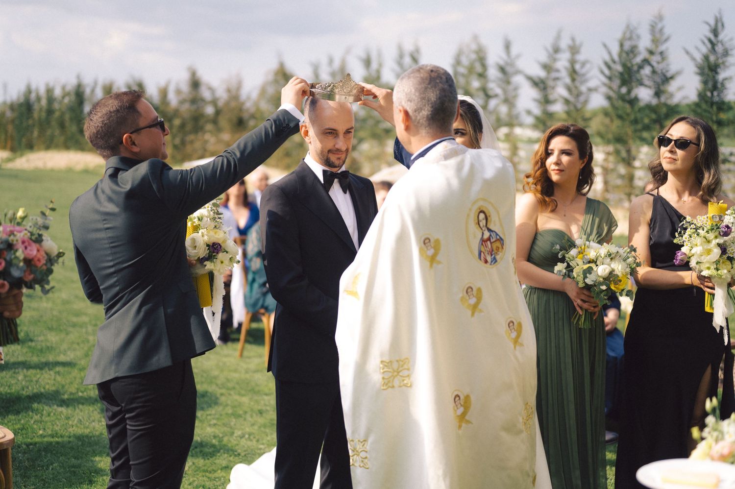 An outdoor wedding ceremony takes place in a field with evergreen trees as religious officiants bless the couple.