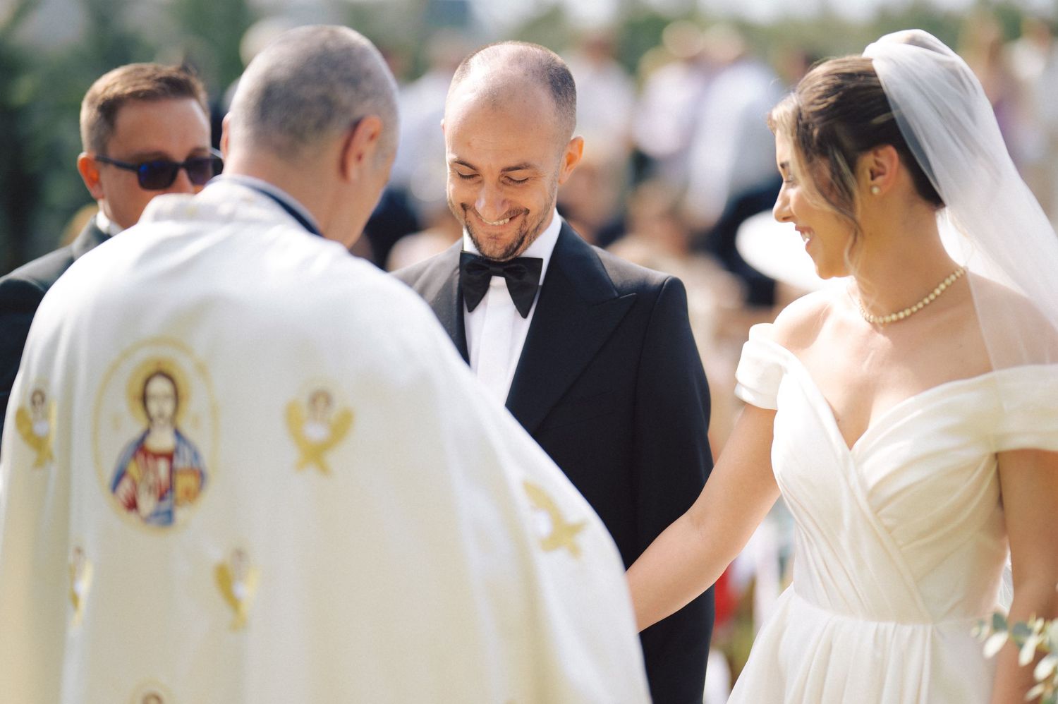 A happy couple exchanges vows with an Orthodox priest during their outdoor wedding ceremony.