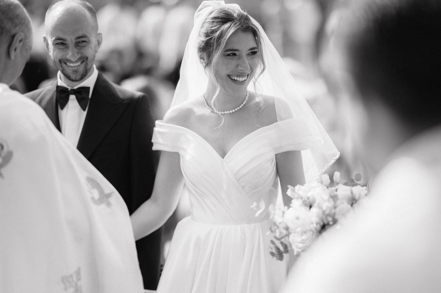 A bride in an elegant off-shoulder white wedding gown smiles while holding a bouquet during her wedding ceremony.