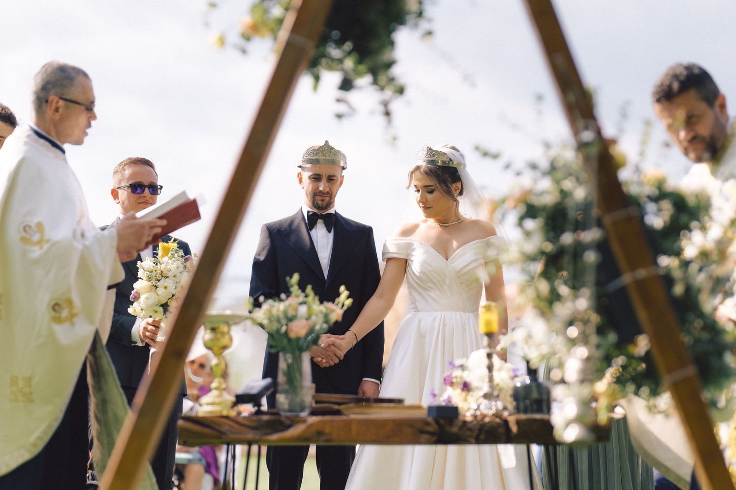 An outdoor wedding ceremony takes place under a geometric wooden arch with natural decor and candlelight.