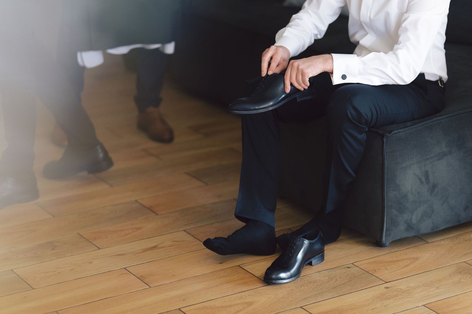Person wearing black dress pants and white shirt sits on ottoman while putting on formal black shoes on hardwood floor.
